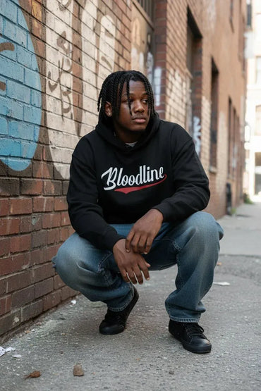A man crouching on a city sidewalk against a brick wall, wearing a black hoodie with 'Bloodline' printed in white script and red underline across the chest, paired with blue jeans and black sneakers.