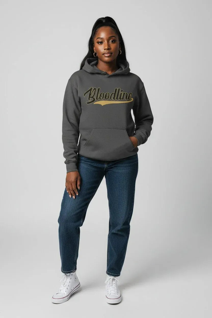 A woman modeling a dark gray hoodie with 'Bloodline' printed across the chest in a metallic gold script, paired with blue jeans and white sneakers, against a light gray studio background.