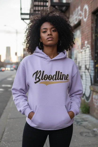 A woman with curly black hair stands on a city sidewalk wearing a light lavender-purple hoodie with 'Bloodline' printed across the chest in black script with a gold underline accent. The background shows an urban street scene with graffiti on a brick wall.
