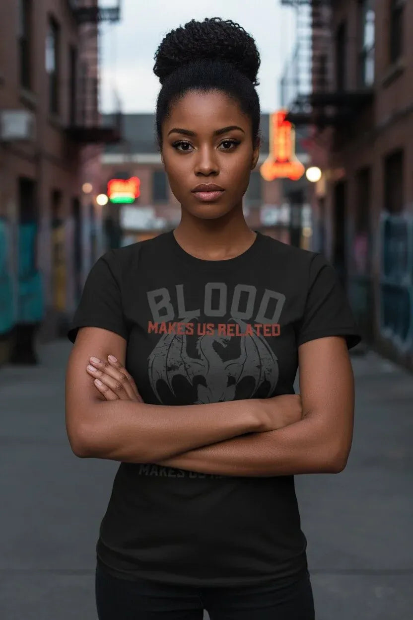 A woman with her hair in a high bun stands on a city street wearing a black graphic t-shirt with the text 'BLOOD MAKES US RELATED' and a gray bat-like emblem.