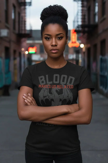 A woman with her hair in a high bun stands on a city street wearing a black graphic t-shirt with the text 'BLOOD MAKES US RELATED' and a gray bat-like emblem.