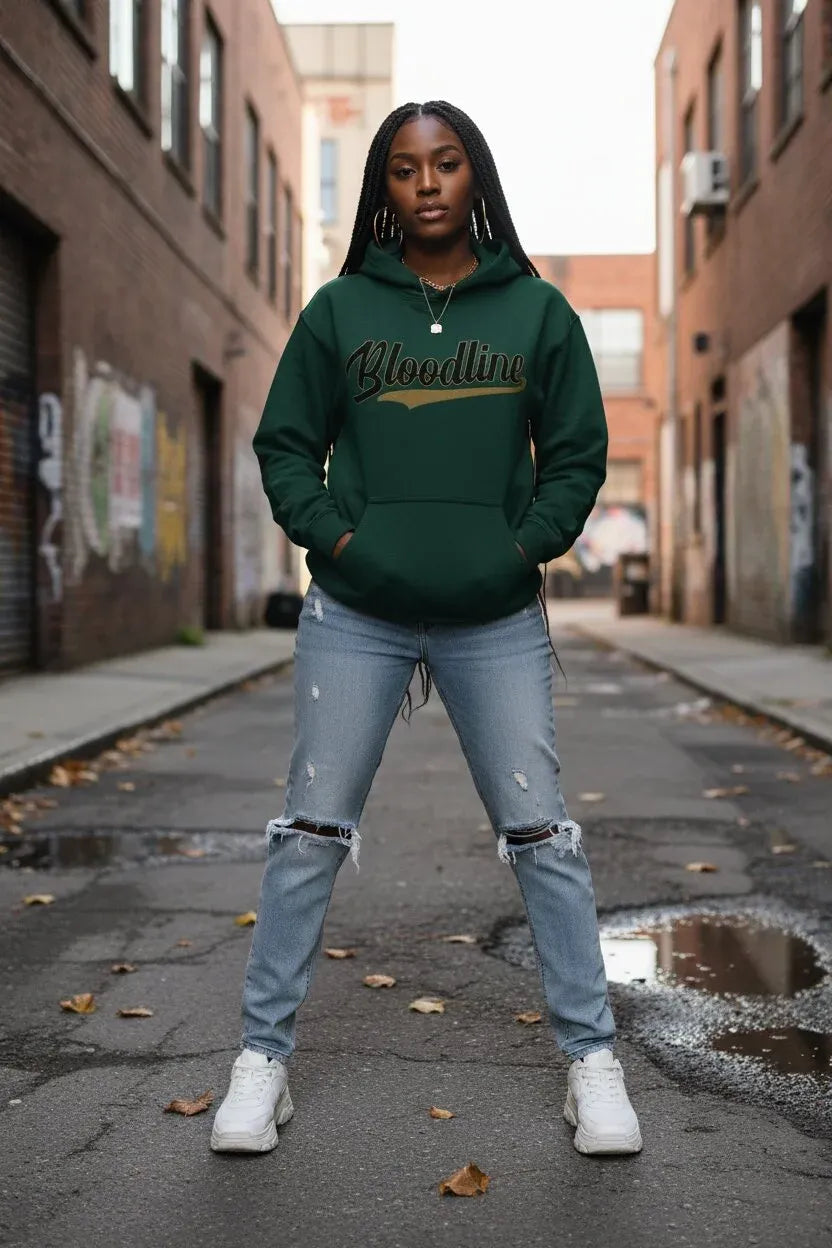 A woman stands in an urban alley wearing a dark green hoodie with 'Blindlin'' printed across the chest in gold and black script, paired with light blue ripped jeans and white sneakers.