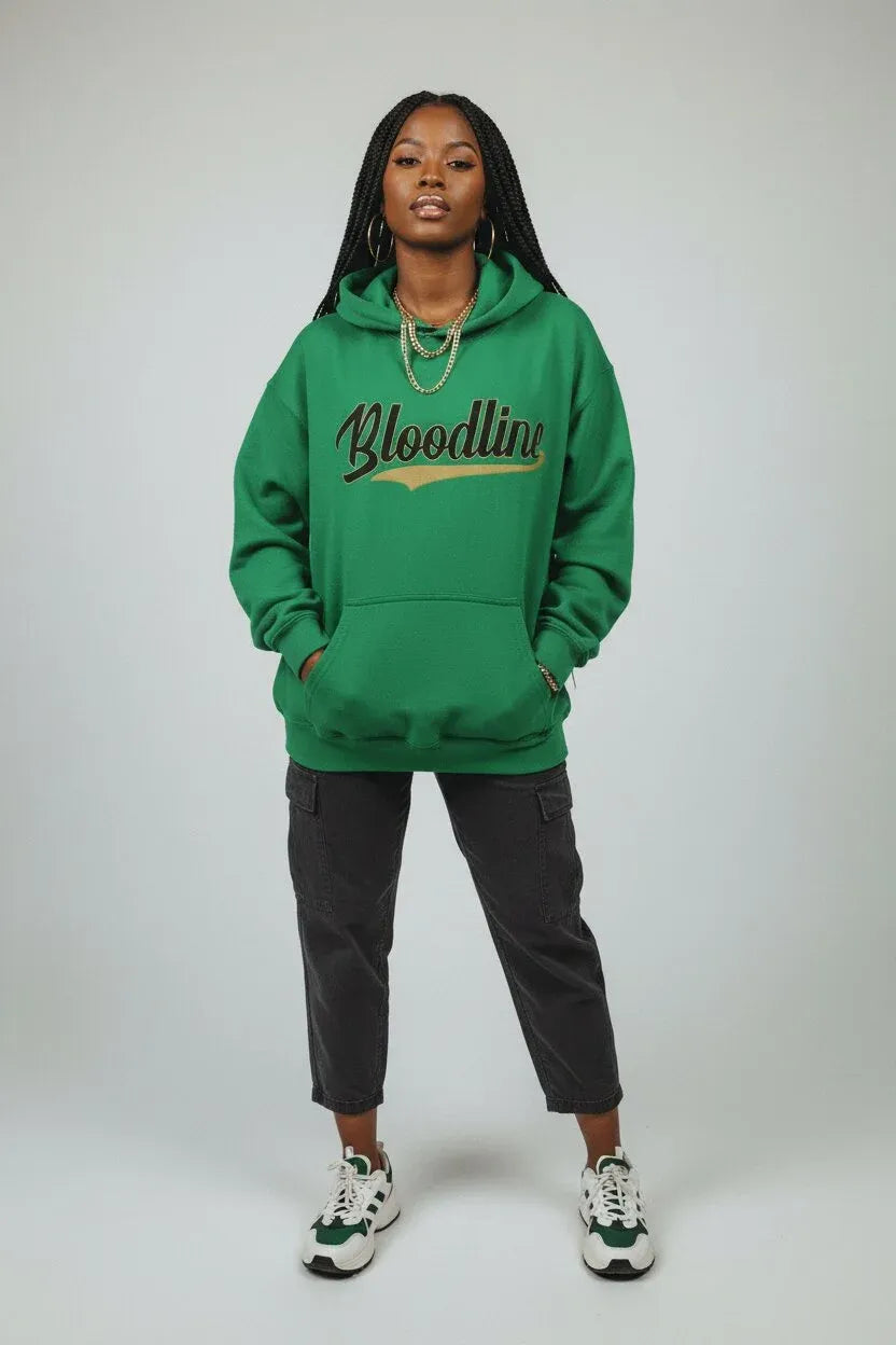 A woman modeling a green pullover hoodie with 'Bloodline' printed across the chest in black script with a gold outline, paired with dark gray pants and white sneakers, against a light gray studio background.