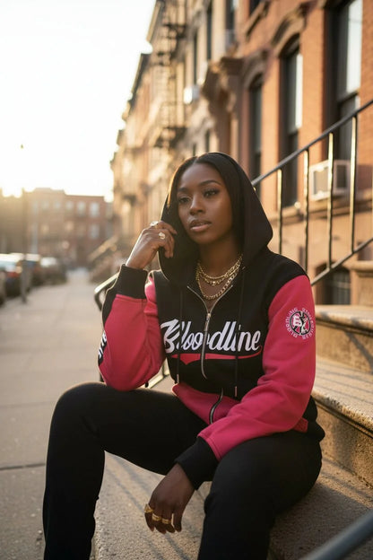 Woman wearing a pink and black jacket with 'Bloodline' branding, sitting on a stoop.