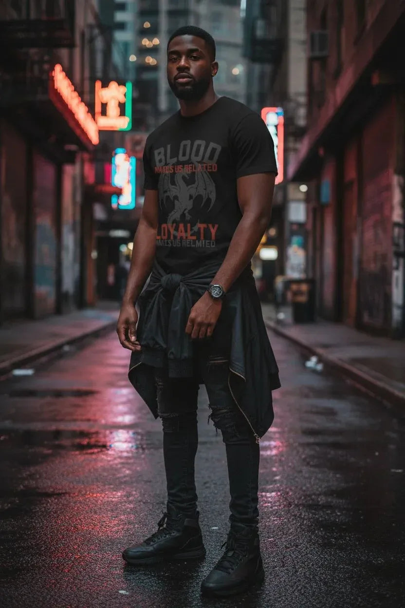 A man stands on a wet city street at night wearing a black graphic T-shirt with distressed text reading 'BLOOD LUST' and 'LOYALTY' in orange, along with black pants and boots. Neon signs and blurred storefront lights appear in the background.