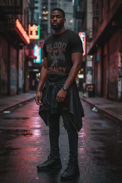 A man stands on a wet city street at night wearing a black graphic T-shirt with distressed text reading 'BLOOD LUST' and 'LOYALTY' in orange, along with black pants and boots. Neon signs and blurred storefront lights appear in the background.