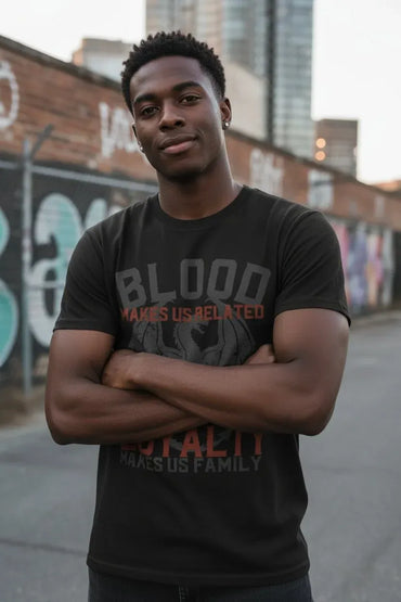 A man wearing a black graphic t-shirt with 'BLOOD' and 'FAMILY' printed on the front, standing with arms crossed in an outdoor urban alleyway.