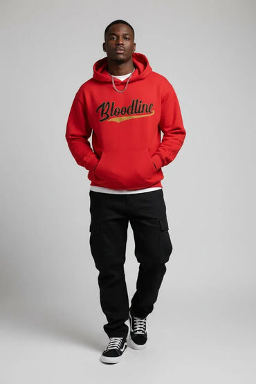 A man modeling a bright red pullover hoodie with 'Bloodline' printed across the chest in black script with a gold outline, paired with black cargo pants and black sneakers, shot against a neutral gray studio background.