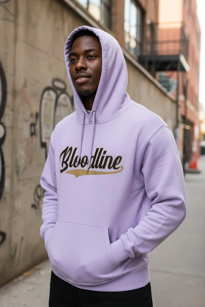 A man wearing a light purple/lavender pullover hoodie with 'Bloodline' printed across the chest in black script with a gold underline, standing on an urban street.