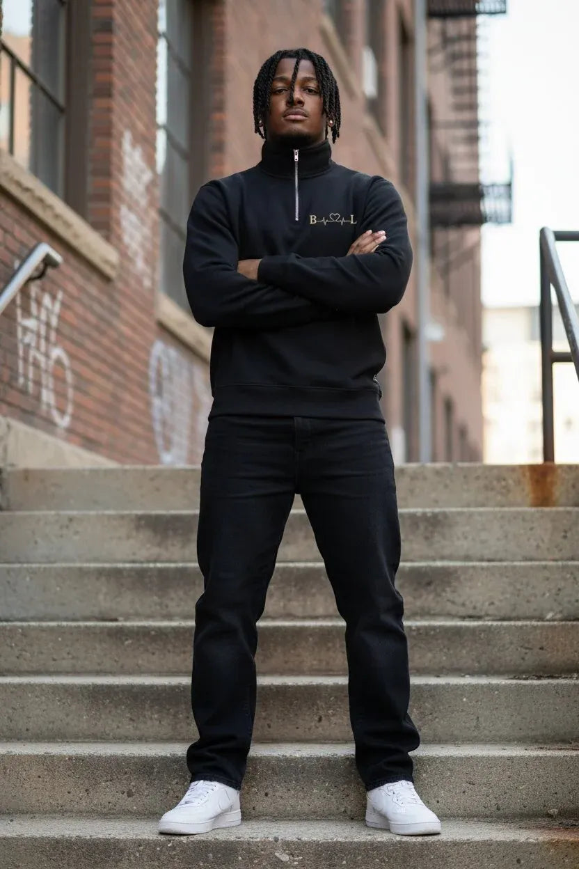 A man stands on concrete stairs wearing a black half-zip sweatshirt with a small gold embroidered logo on the chest, black sweatpants, and white sneakers. The background shows brick buildings and outdoor city architecture.