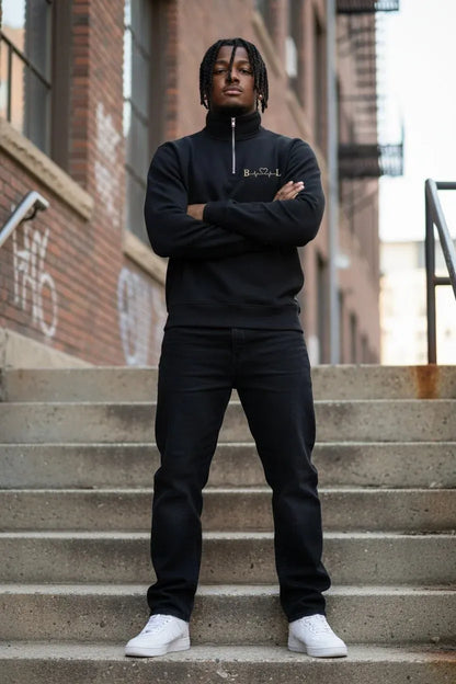 A man stands on concrete stairs wearing a black half-zip sweatshirt with a small gold embroidered logo on the chest, black sweatpants, and white sneakers. The background shows brick buildings and outdoor city architecture.