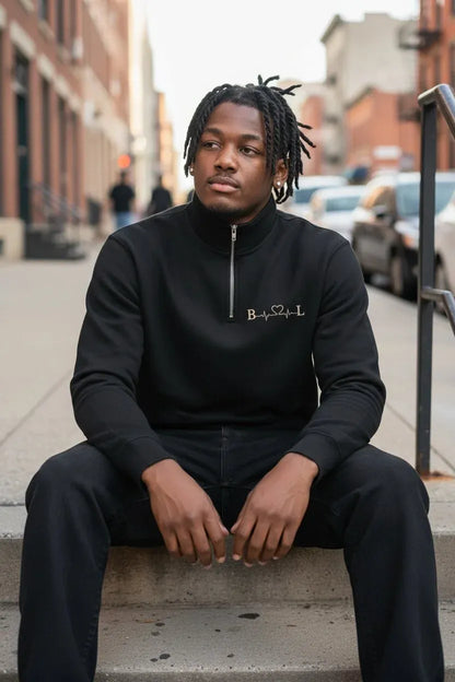 A man with dreadlocks sits on concrete steps on a city street, wearing a black long-sleeve quarter-zip sweatshirt with a small white embroidered logo on the chest and black pants.