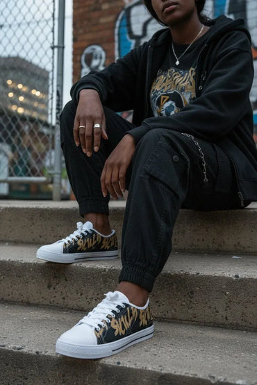 A person sitting on concrete steps wearing black low-top sneakers with a black, gold, and white graphic pattern on the sides, paired with a black graphic t-shirt and black hoodie.