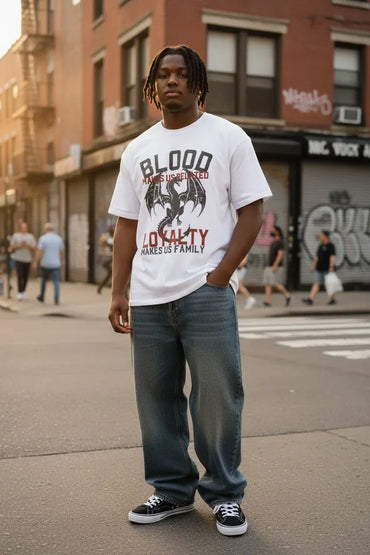 A man standing on a city street wearing a white graphic t-shirt with a red and black dragon design and the text 'BLOOD - LOYALTY, paired with blue jeans and black sneakers.