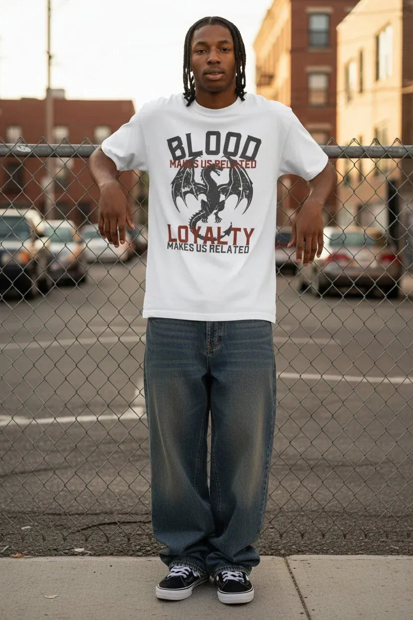 A man stands in front of a chain-link fence wearing a white graphic t-shirt with a black dragon design and the words 'BLOOD - LOYALTY' in red and black, paired with loose blue jeans and black sneakers.