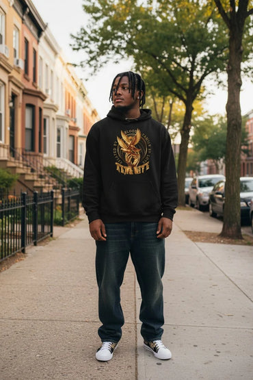 A man stands on a city sidewalk wearing a black hoodie with a large gold graphic on the front, paired with dark blue jeans and white sneakers. The background shows a residential street with brown townhouses, trees, and parked cars.