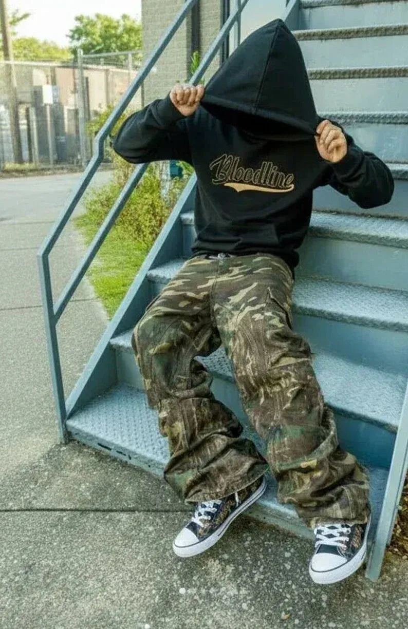 A person sitting on outdoor metal stairs wearing a black hoodie with a gold script logo on the chest, camouflage cargo pants, and black-and-white sneakers.
