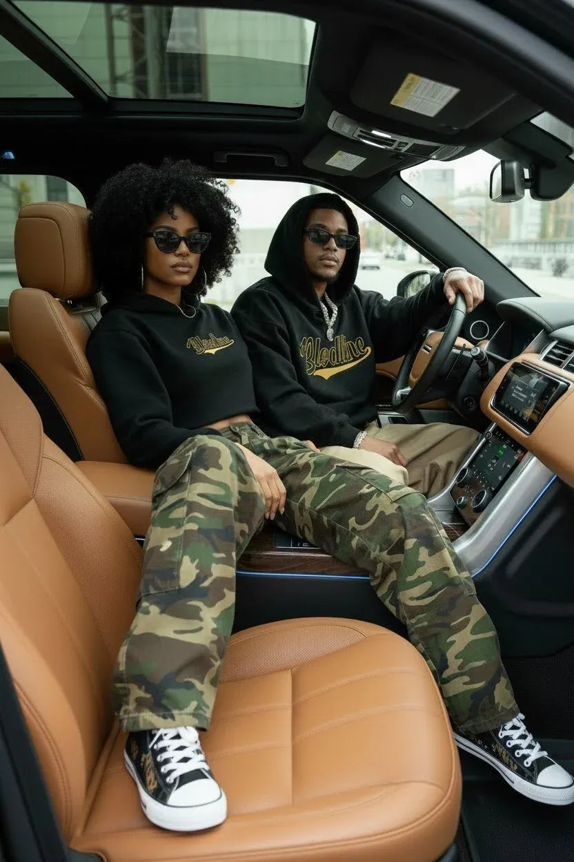 Two models sitting inside a luxury car with tan leather seats. The model on the left wears a black graphic hoodie and camouflage pants, while the model on the right wears a black hoodie and khaki pants.