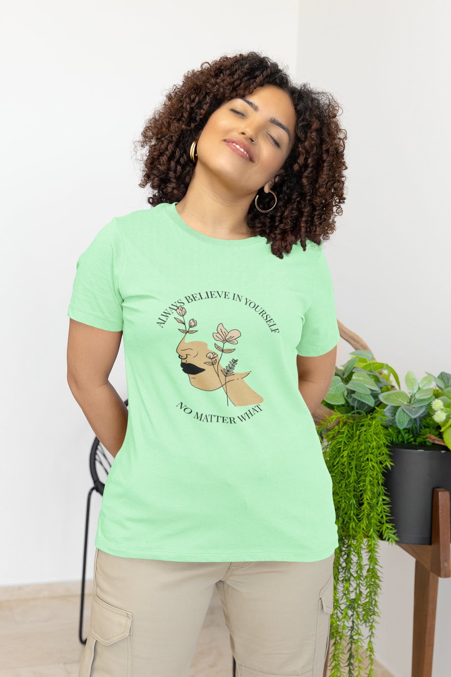 Woman wearing a light green t-shirt with a motivational quote and dog illustration, standing in a room with plants.