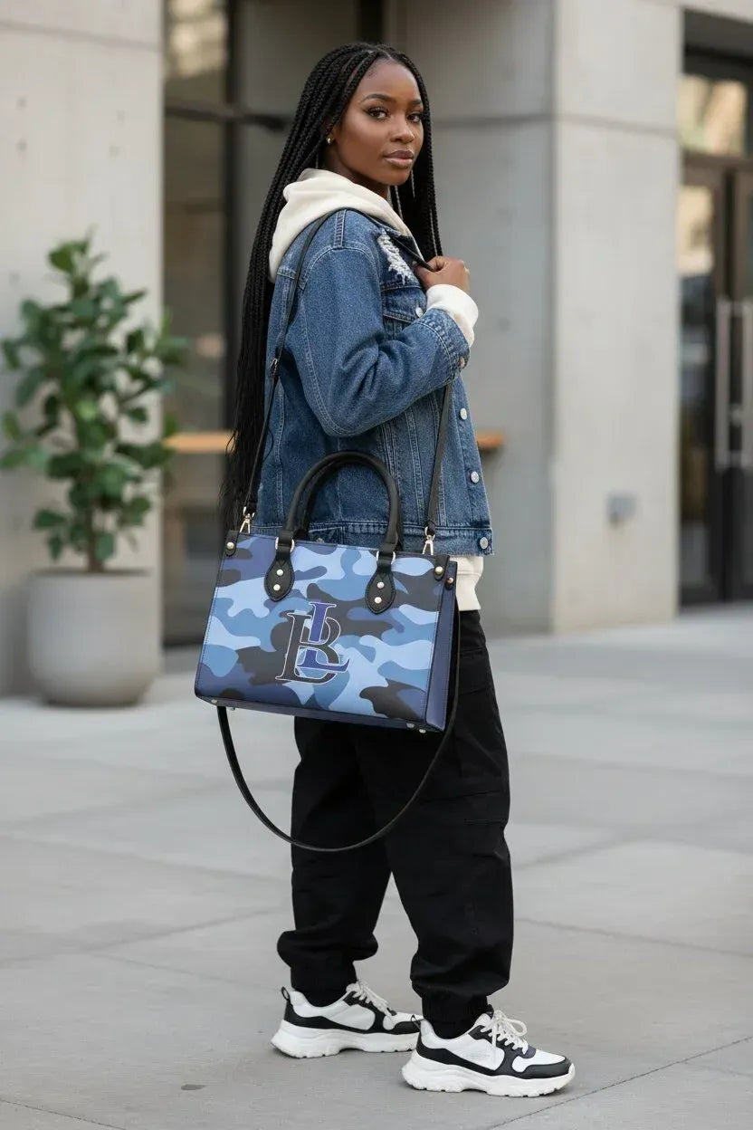 A woman wearing a blue denim jacket, black sweatpants, and white sneakers carries a blue camouflage tote bag with a white monogram logo, standing outdoors on a paved walkway.