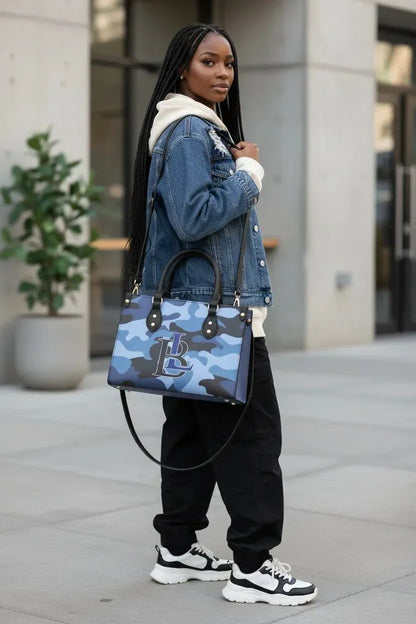 A woman wearing a blue denim jacket, black sweatpants, and white sneakers carries a blue camouflage tote bag with a white monogram logo, standing outdoors on a paved walkway.