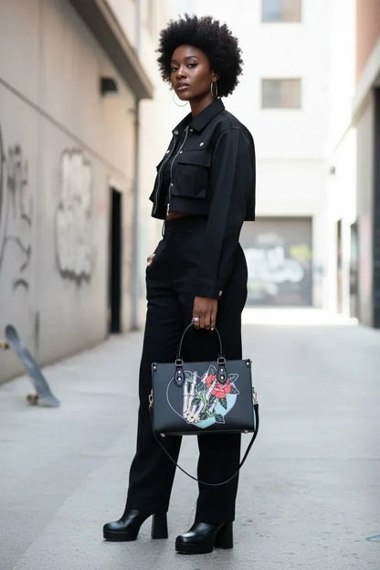A woman stands on a city sidewalk wearing a black cropped jacket and matching black pants, holding a black structured handbag with colorful graffiti-style artwork. The background shows an urban alley with buildings and a skateboard.