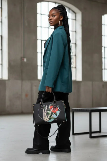A woman in a teal blazer and black pants stands in a concrete industrial loft, holding a black leather handbag with a skeleton hand graphic and a red rose detail.