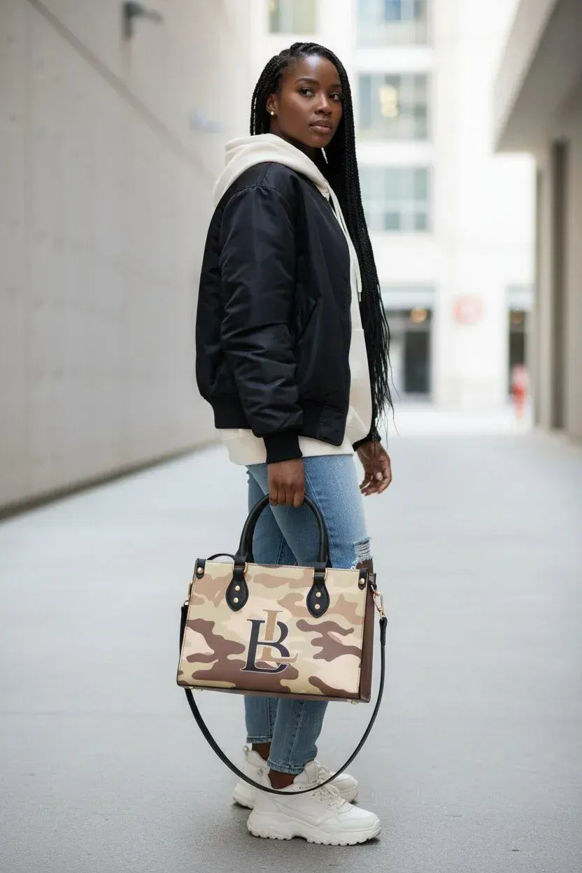 A woman stands in a city alleyway holding a beige and brown camouflage tote bag with a large 'B' logo on the front. She is wearing a black and white puffer jacket, a white hoodie, ripped blue jeans, and white sneakers.
