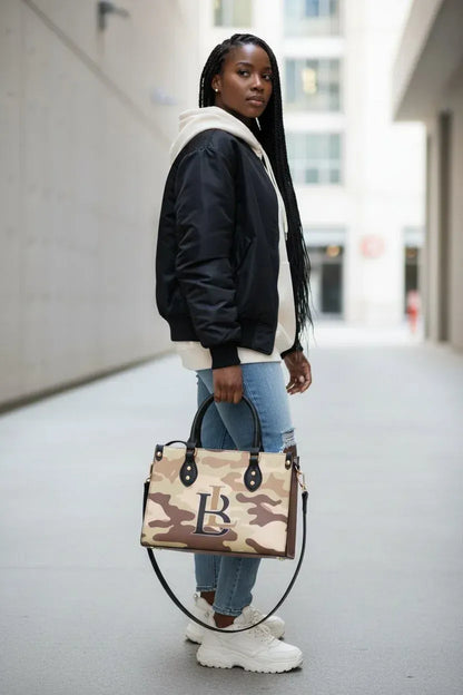 A woman stands in a city alleyway holding a beige and brown camouflage tote bag with a large 'B' logo on the front. She is wearing a black and white puffer jacket, a white hoodie, ripped blue jeans, and white sneakers.
