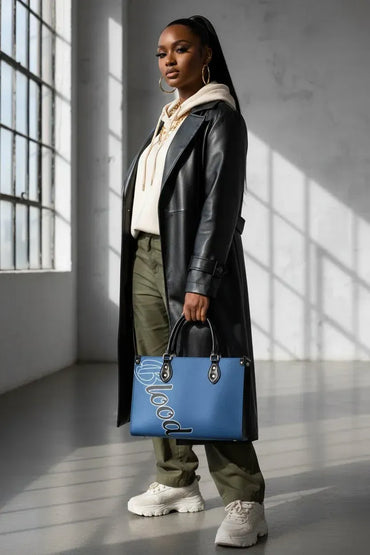 A model stands in a sunlit industrial loft holding a blue leather tote bag with white script lettering. She is wearing a black leather trench coat, a cream hoodie, olive green pants, and white sneakers.