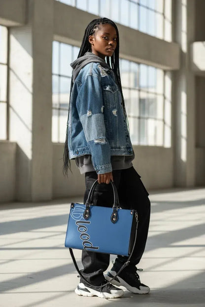 A woman stands in a sunlit industrial interior holding a bright blue tote bag with large white script lettering. She wears a distressed blue denim jacket over a gray hoodie, black pants, and white sneakers.