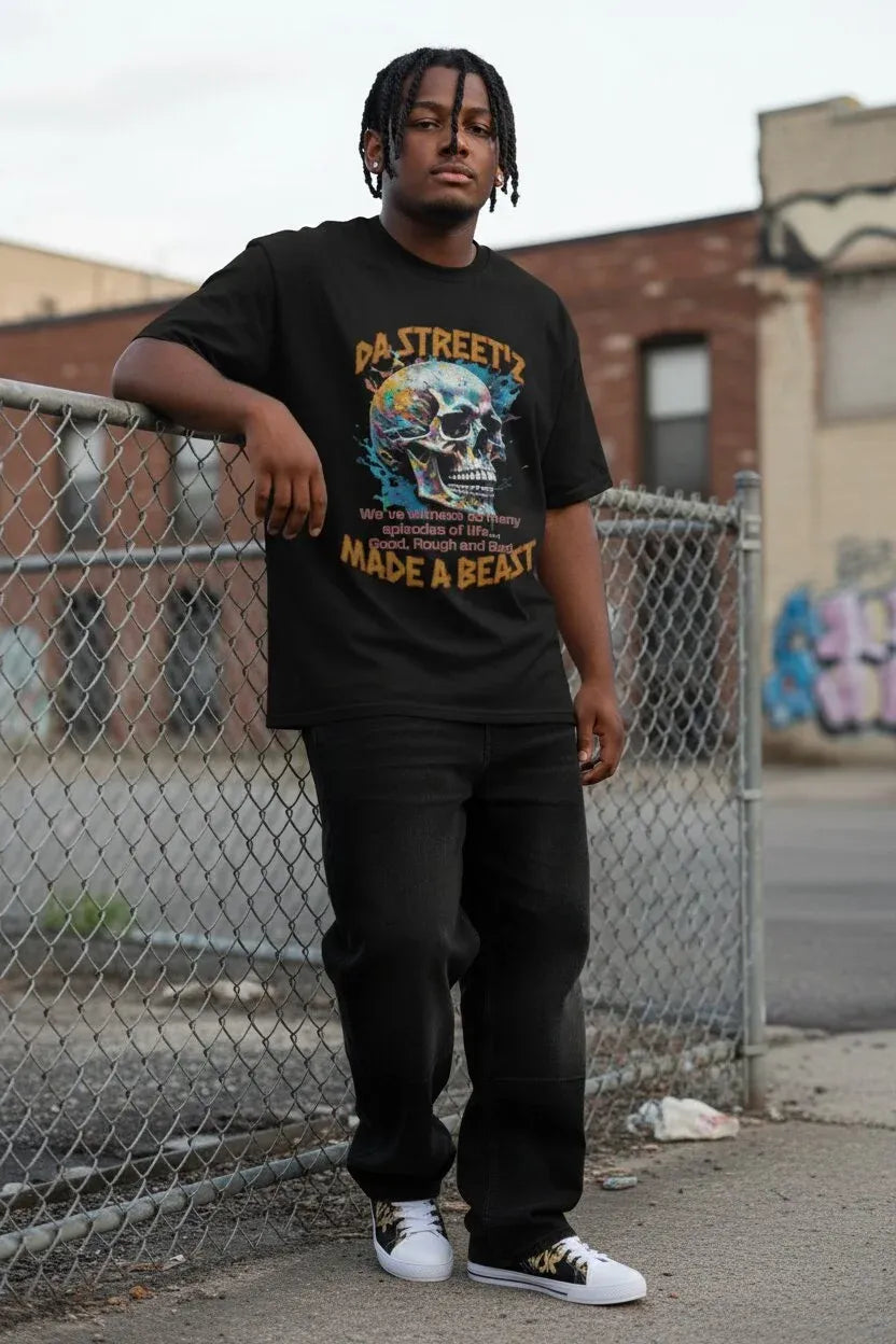 A man wearing a black graphic t-shirt with a skull design and the text ' MADE A BEAST', paired with black sweatpants and white sneakers, standing in front of a chain-link fence in an urban outdoor setting.