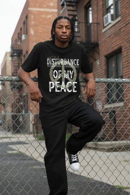 A man on a city street fence wearing a black graphic t-shirt with the text 'DISTURBANCE OF MY PEACE' 