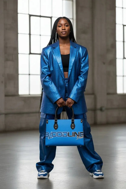 A model stands in a warehouse setting wearing a shiny metallic blue blazer and matching pants, holding a blue tote bag with the brand name 'XOODLINE' printed on it.
