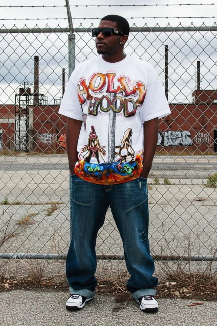 Person wearing a colorful 'Hollywood' t-shirt with graphic designs, standing in front of a chain-link fence.