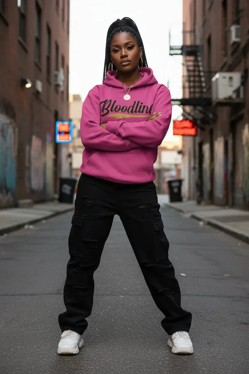 A woman stands in an urban alley wearing a bright pink hoodie with 'Bloodline' printed across the chest in black and gold, paired with black cargo pants and white sneakers.