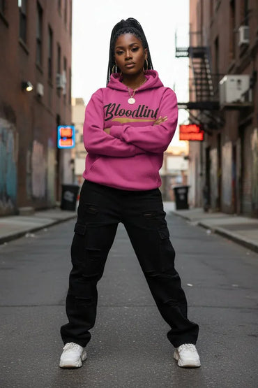 A woman stands in an urban alley wearing a bright pink hoodie with 'Bloodline' printed across the chest in black and gold, paired with black cargo pants and white sneakers.