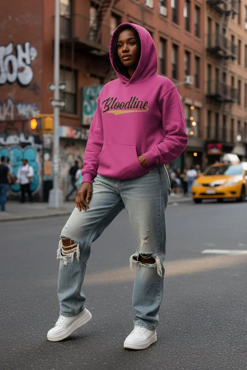 A woman stands on a city street wearing a bright pink hoodie with 'Bloodline' printed across the chest in black and gold, light-wash ripped jeans, and white sneakers.
