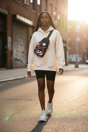 A woman walking down a sunlit city street wearing a cream hoodie, black biker shorts, white sneakers, and a red, black, and white patterned crossbody sling bag.
