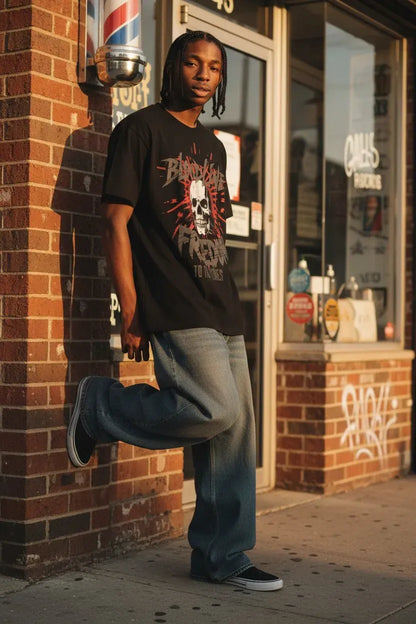 A man stands on a city sidewalk in front of a brick storefront, wearing a black graphic T-shirt with a skull print, loose blue jeans, and black sneakers.