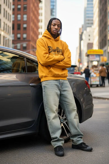 A man wearing a mustard yellow hoodie with 'Bloomin" printed in black across the chest, paired with light-wash jeans and black sneakers, standing next to a dark gray car on a city street.
