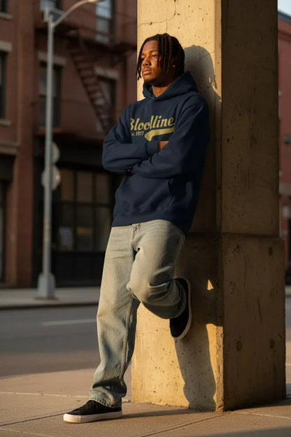 A man wearing a navy blue hoodie with 'Bloclines' in yellow lettering, paired with light-wash jeans and black sneakers, leaning against a concrete pillar on a city street.