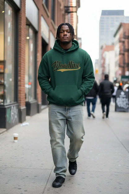 A man walking on a city sidewalk wearing a dark green hoodie with 'Bloodline' printed across the chest in gold and white script, paired with light gray jeans and black shoes.