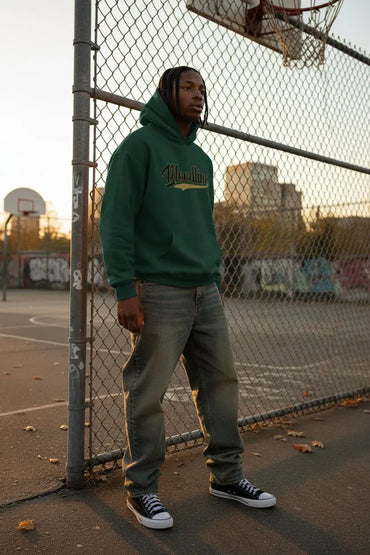 A man wearing a forest green hoodie with a gold and white graphic on the chest, paired with gray jeans and black sneakers, standing on an outdoor basketball court behind a chain-link fence.