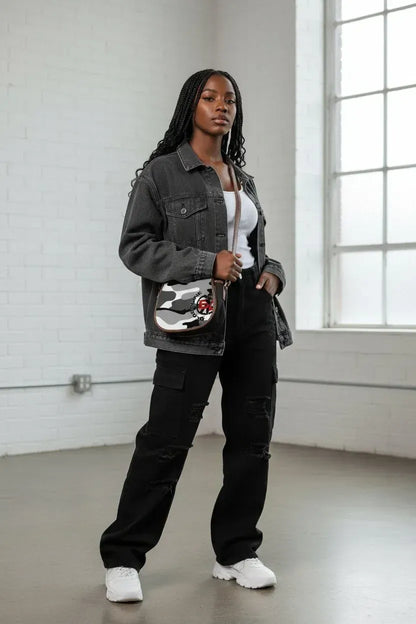 A woman stands in a bright white brick room wearing a dark gray denim jacket, black cargo pants, and white sneakers, while carrying a small silver and black crossbody bag with a red and white graphic.