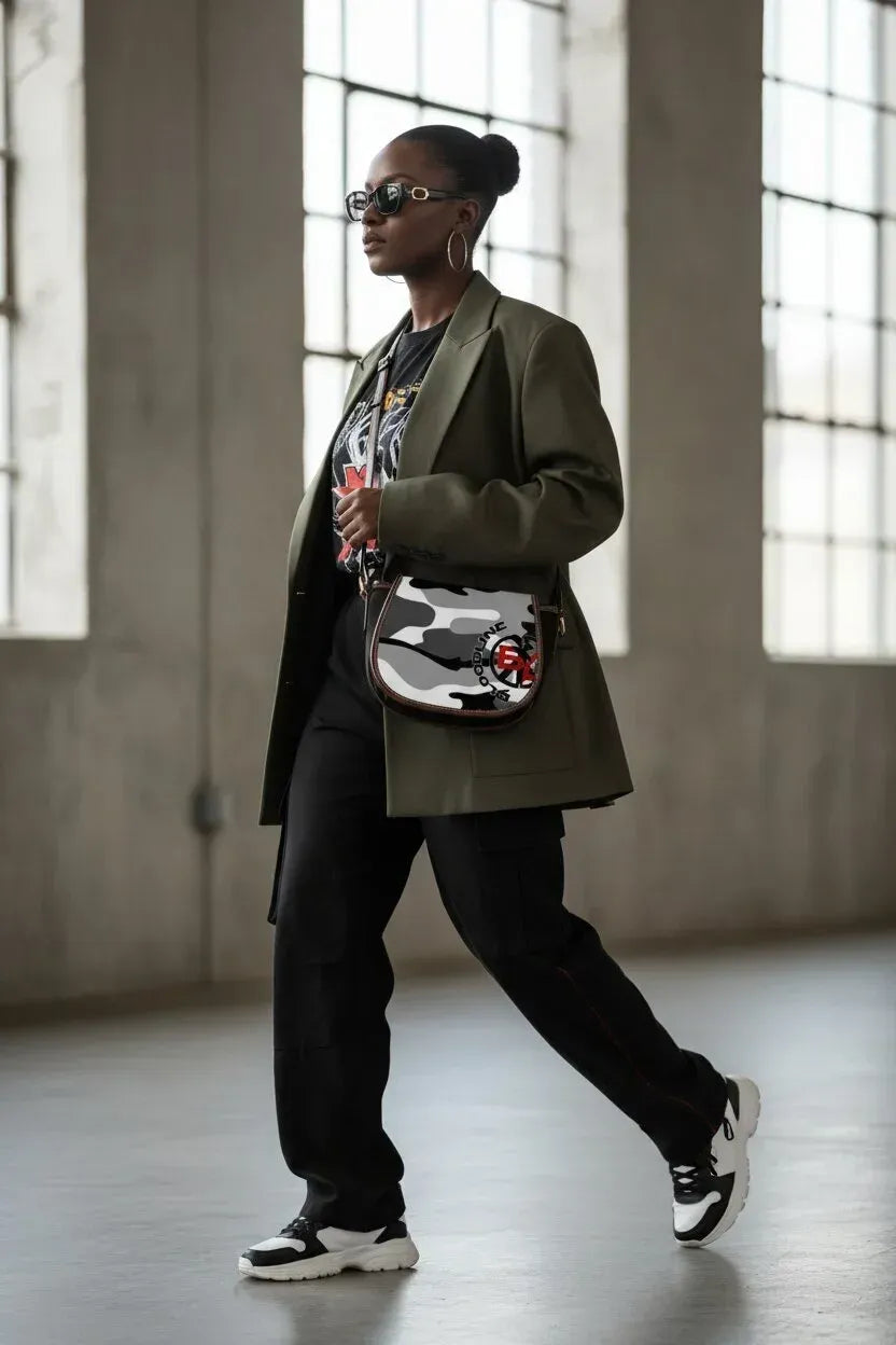 A woman walking in a large industrial loft with high windows, wearing a dark olive green blazer, black graphic t-shirt, black sweatpants, and a black, white, and gray camouflage patterned crossbody bag.