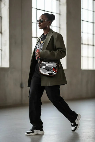 A woman walking in a large industrial loft with high windows, wearing a dark olive green blazer, black graphic t-shirt, black sweatpants, and a black, white, and gray camouflage patterned crossbody bag.