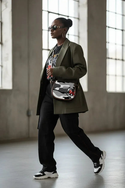 A woman walking in a large industrial loft with high windows, wearing a dark olive green blazer, black graphic t-shirt, black sweatpants, and a black, white, and gray camouflage patterned crossbody bag.