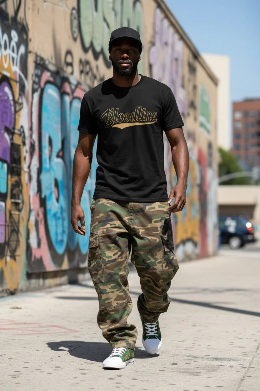 A man walking outdoors on a city sidewalk wearing a black t-shirt with a gold 'Bloodline' graphic, camouflage cargo pants, and green and white sneakers, set against a graffiti-covered wall.