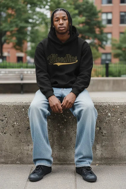 A man sitting on a concrete ledge outdoors wearing a black hoodie with a gold 'Bloodline' graphic, light blue jeans, and black sneakers.